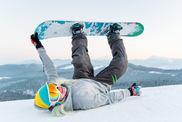 Excited young snowboarder woman having fun on the slope, lying on the snow with her legs in the air...