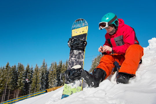 Young male snowboarder sitting on the snow, preparing for riding downhill on a beautiful sunny winter day. copyspace ski resort recreation sports activity hobby leisure concept