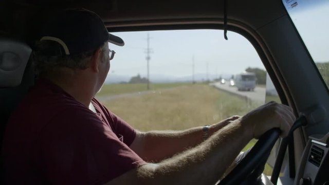Profile view of truck driver wearing burgundy T-shirt, glasses and cap, waiting to make a turn at a busy rural intersection on a warm summer day. Interior, hand-held slow motion 4K recorded at 60fps.