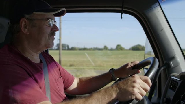 Profile view of man wearing burgundy T-shirt, glasses and cap, driving truck past farmland on a warm summer day. Interior, hand-held slow motion 4K recorded at 60fps.