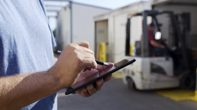 Man's hands making notes on electronic tablet while forklift driver loads a truck with pallets in background. Close up view with focus on hands, 4K