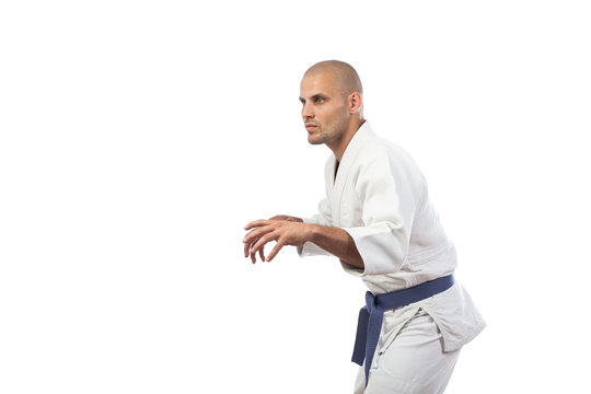 A young athletic man is a fighter in a white kimono for judo, jiu jitsu, sambo with a blue belt standing in a fighting rack on a white insulated background