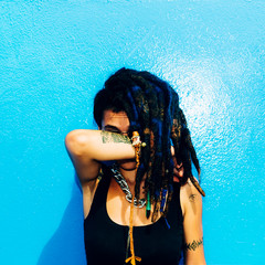 Rasta Latino Girl with dreadlocks, piercings, tattoos and stylish accessories on the blue wall.