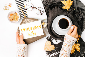 Woman holding card with word I Love Autumn. Workspace with laptop,  coffee cup wrapped in scarf,   glasses. Stylish office desk. Autumn  concept.  Flat lay, top view
