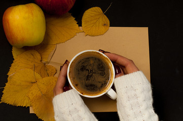 Woman's hand in a sweater holds a cup of coffee on the background of an envelope with fried leaves and colored apples, an autumnal mood