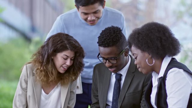 Tilt up of diverse business team sitting outdoors on bench, watching something on tablet, smiling and discussing it