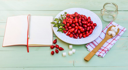 Fresh red cornel berries on white plate, preparing for homemade cornelian cherry jam, surrounded by jelly jar, flax napkin, vintage spoon, sugar, spices,notebook for recipes, rustic wooden background.