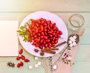 Fresh red cornel berries on white plate, preparing for homemade cornelian cherry jam, surrounded by jelly jar, flax napkin, vintage spoon, sugar, spices,notebook for recipes, rustic wooden background.