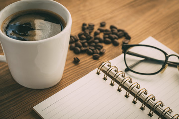 White Cup of coffee, eyeglasses, sketchbook notebook  or notepad, with coffee beans on brown wooden table background in coffee shop