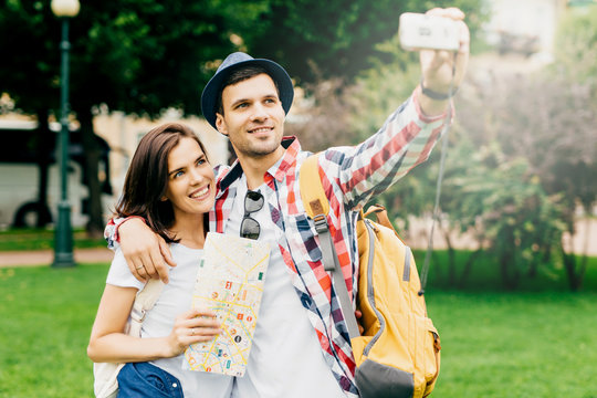 Handsome guy embracing his beautiful girlfriend, travelling abroad, using map for finding right destination and places, making photo of themselves, having cheerful expressions. Tourism concept