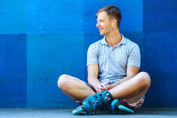 Happy handsome young roller in inline skates sitting against blue wall.