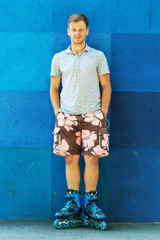 Happy handsome young roller in inline skates sitting against blue wall.