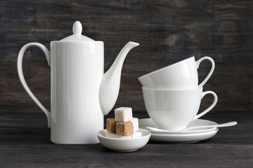 White porcelain coffee pot, stacked cups and sugar cubes on wooden rustic table.