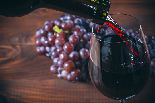 Pouring red wine into the glass with a bunch of red grapes against wooden background