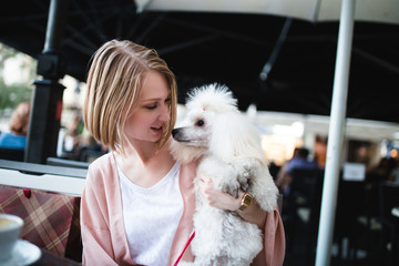 Beautiful and fashionable young woman with shopping bags and white dwarf poodle sitting in cafe bar...