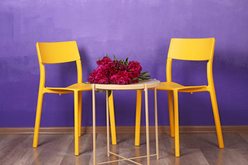 Chairs and table with peony flowers near lilac wall