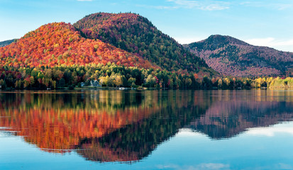 The hills covered with red maple forests are reflected in a lake in Quebec on a beautiful autumn evening - obrazy, fototapety, plakaty