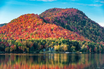 Fototapeta premium Wzgórza pokryte czerwonymi lasami klonowymi za drewnianym domem nad brzegiem jeziora w Quebecu, w piękny jesienny wieczór.