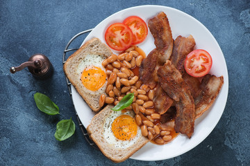 English breakfast with fried bacon, eggs in the basket, beans and tomatoes. Flat-lay on a blue stone background