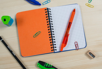 Top view of closed orange notebook with pen, paper clips and  pencil sharpener on wooden background. School stationery, office and student accessories.