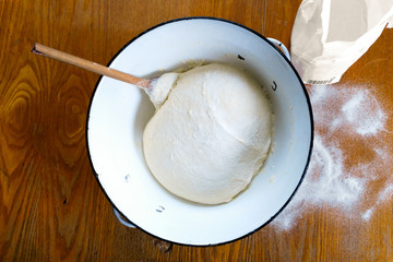 Rising yeast dough with wooden spoon in white enamel metal bowl on wooden table from above. Spilled flour.