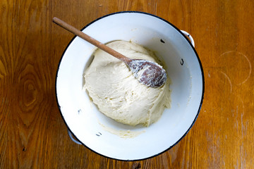 Unleavened yeast dough with wooden spoon in white enamel metal bowl on wooden table from above.