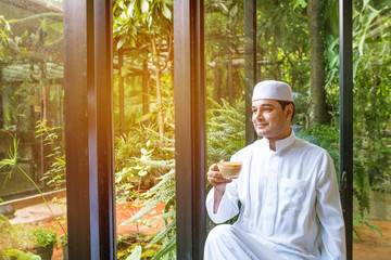 Arab muslim man in white dress hand hold glass of late coffee,eyes look forward and sit in coffee shop.