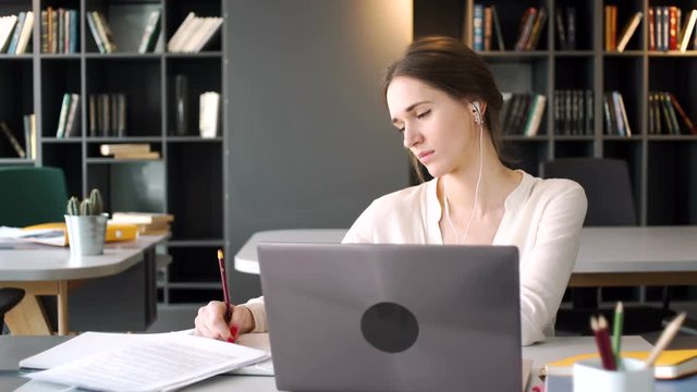 young beautiful woman during video conference call and webinar education listen and make notes in notebook looking at laptop screen sitting at the table in co working office