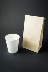 White paper cup for tea, coffee or juice presented near blank recycled paper bag, isolated on black background. Space for text, mock up.