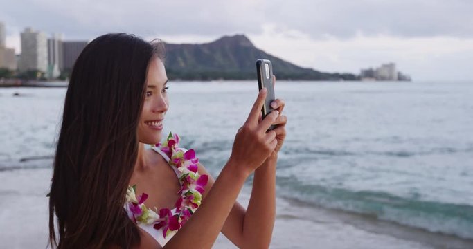 Smiling young woman photographing through smart phone at Waikiki Beach. Happy female tourist is wearing orchid lei garland during vacation at island in Honolulu using mobile cell phone.