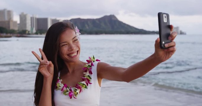 Happy young woman taking selfie using smartphone at Waikiki Beach. Girl tourist is wearing orchid lei garland on vacation gesturing peace sign photographing using mobile phone app. Oahu, Hawaii.