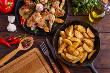 Grilled chicken wings, baked potatoes, herbs and spices on kitchen table. Hot fresh appetizing countryside food. Cooking concept, meat dishes, homemade cuisine