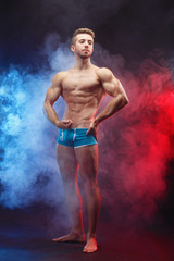 Young handsome ripped muscular man bodybuilder with perfect abs, shoulders,biceps and triceps demonstrating huge muscles standing in smoke, full length portrait