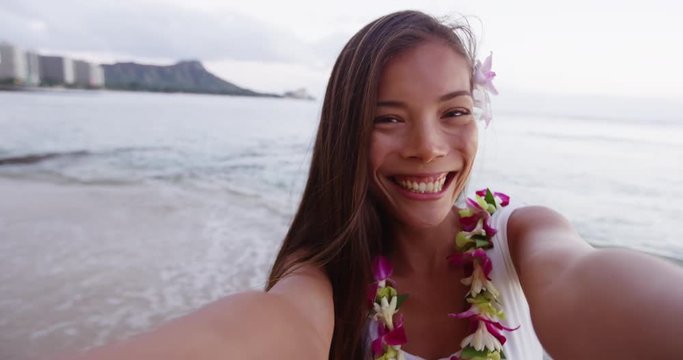 Cheerful young woman taking selfie at Waikiki Beach. Smiling beautiful female tourist is wearing orchid lei garland during vacation. She is puckering lips blowing kiss at camera in Honolulu.