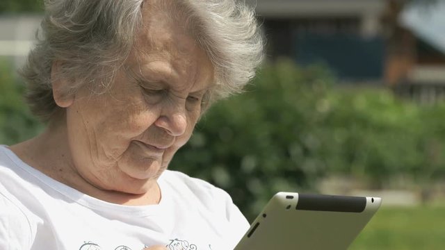 Mature elderly woman dressed t-shirt sitting on the background the green park outdoors in summer holds a silver portable computer tablet for looking the images. Slow motion