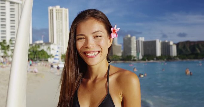 Happy young woman with surfboard at Waikiki Beach. Beautiful female surfer tourist is on summer vacation at island in Honolulu. She is wearing bikini top against buildings and sky.