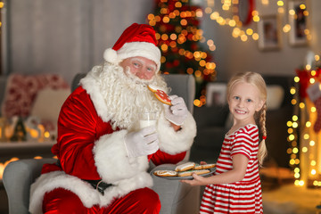 Fototapeta na wymiar Little girl treating Santa with tasty cookies and milk in room with beautiful Christmas decorations