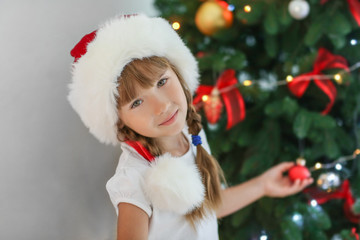 Cute little girl in Santa hat decorating Christmas tree at home