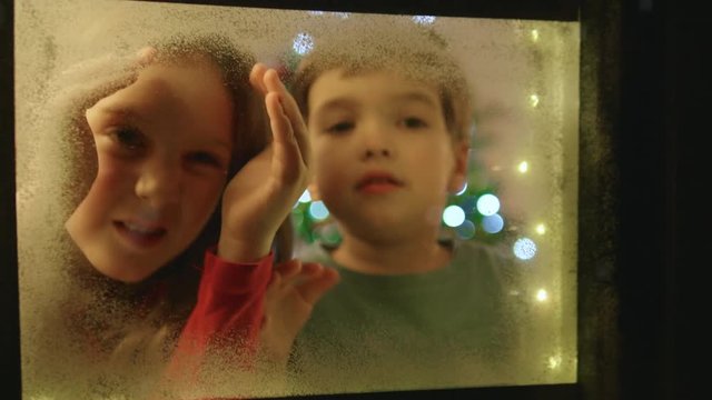 Adorable little boy and girl looking through the window and admiring first snow flakes and fireworks on Christmas evening.
