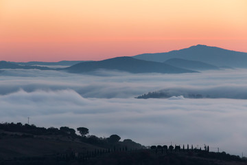 A valley filled by fog at sunset, with some hills and trees in the foreground and other hills and mountains in the foreground