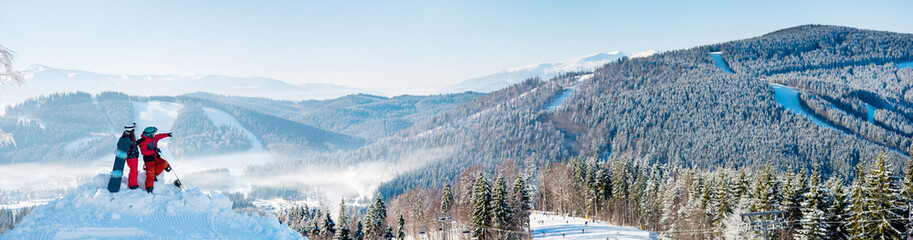 Extra wide panorama of the Carpathians mountains landscape, forests in a white haze, ski slopes, winter ski resort Bukovel. Two snowboarders enjoying, resting on top of the mountain on a sunny day - obrazy, fototapety, plakaty