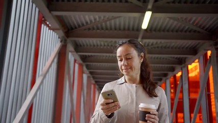 adult brunette is holding smartphone in hand and cup of coffee in other, walking and looking in screen