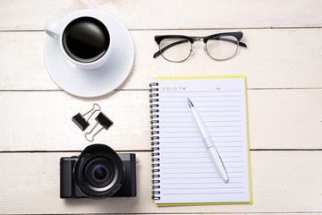 White set. Camera, pen, eyeglasses, coffee and notebook on table. Top view.
