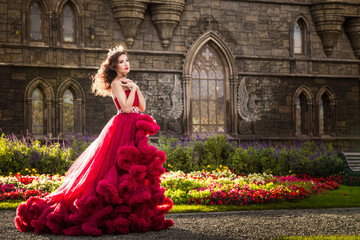 A beautiful woman, a queen in a burgundy lavish dress, walks along a flowering garden. Ancient, Gothic castle on the background. Copy space