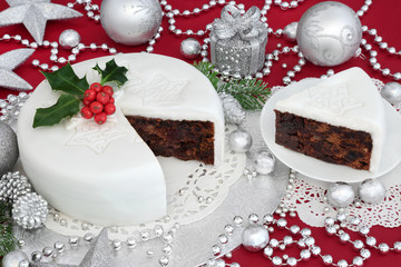 Traditional iced christmas cake and slice with holly, bauble decorations and silver foil wrapped chocolates on red background.
