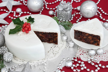 Traditional iced christmas cake and slice with holly, bauble decorations and silver foil wrapped chocolates on red background.

