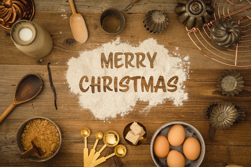 Flatlay collection of tools and ingredients for home baking on a dark wooden table with the words merry christmas written in flour in the center. Shot from above