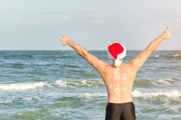 Man in Santa hats with the inscription New Year on the back on the beach. Thumbs up. Back view