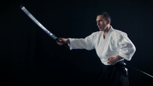 Portrait Shot of the Aikido Master Wearing Traditional Samurai Hakama Clothes Takes His Japanese Sword out of Scabbard and Swings with It. He's in the Spotlight Darkness Surrounds Him. 