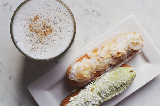 Latte topped with cinnamon and cardamom and two eclairs on marble table